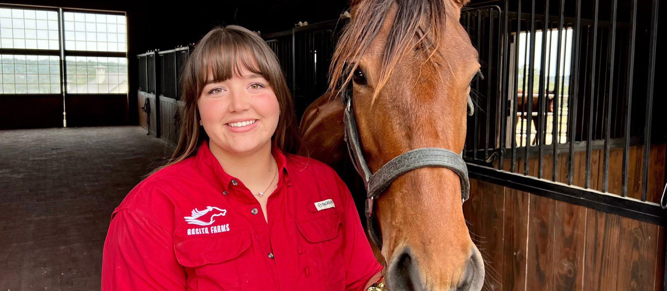 Abby wearing a red rosita farms shirt and posing with a young horse in the stalls.